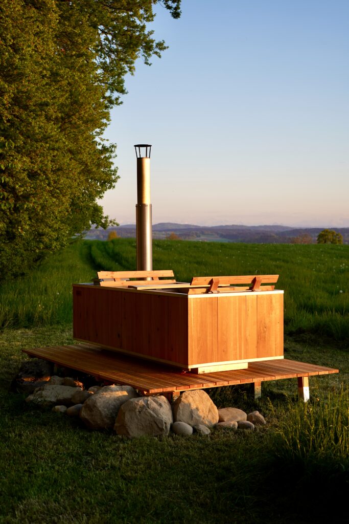 Rechteckiger Holz-Hot-Tub mit integriertem Holzofen und hohem Edelstahlschornstein auf einer Holzplattform in einer blühenden Bergwiese. Große Natursteine stützen die Konstruktion, links ragt ein mächtiger Laubbaum ins Bild, im Hintergrund die schneebedeckten Alpen unter strahlend blauem Himmel.