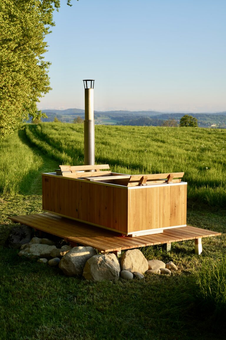 Seitenansicht eines rechteckigen Holz-Hot-Tubs mit Edelstahlschornstein auf Holzplattform in sonniger Alpenwiese mit Bergpanorama.